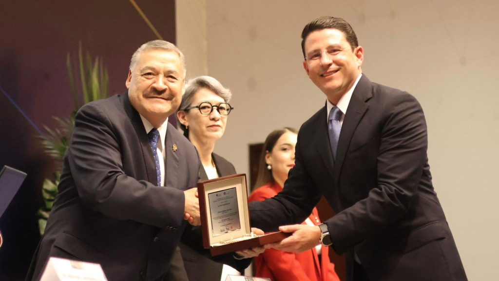 Two Men in Suits Shake Hands As One Presents a Framed Award at a Formal Ceremony, with Onlookers in the Background. › Guadalupa Ciudadana