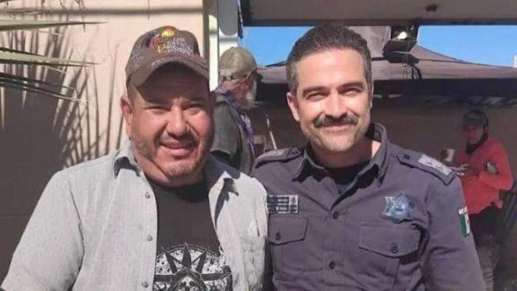 Two Men Pose for a Casual Photo Outdoors; One in a Dark Police-style Uniform with a Mustache, the Other in a Cap and Graphic T-shirt. › Guadalupa Ciudadana