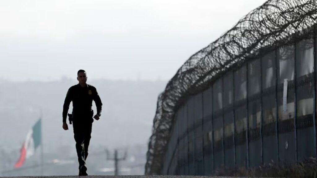 Border Patrol Officer Walking Along a Road Beside a Tall Razor-wire Fence, with a Distant Flag Visible in the Background. › Guadalupa Ciudadana